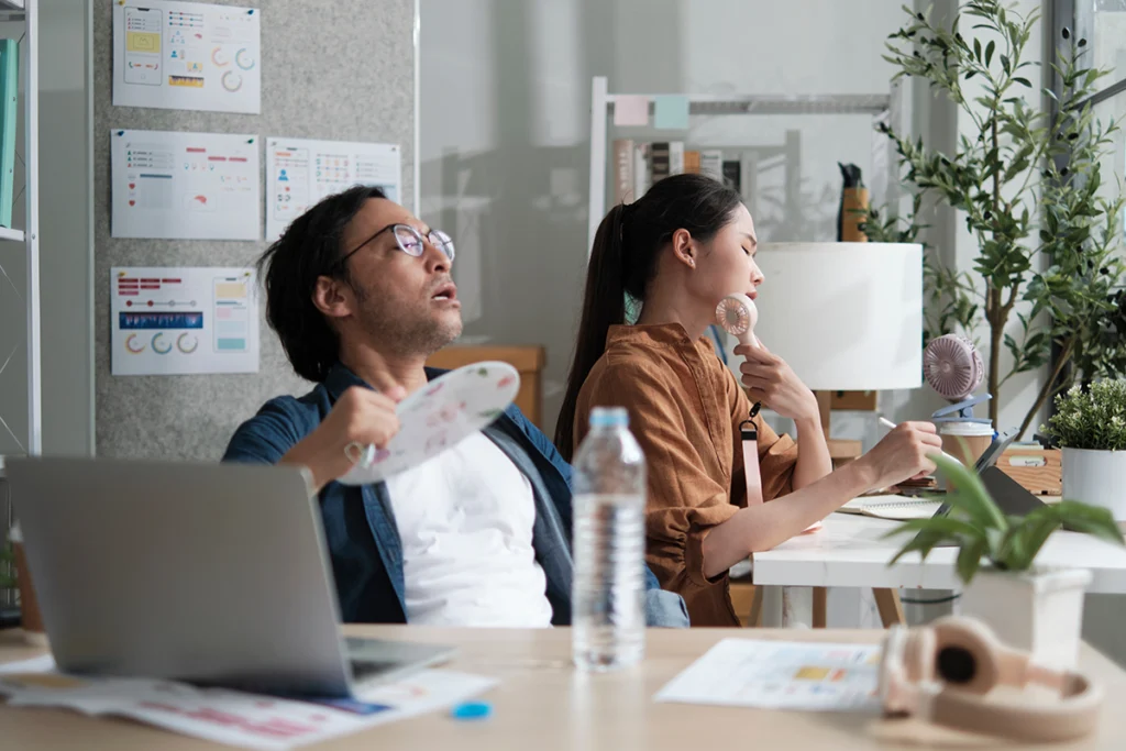 Office workers using handheld fans and looking overheated at their desks in a warm indoor workspace. Best for blogs about offices that are too hot, poor cooling, indoor comfort problems, and commercial AC not keeping up.