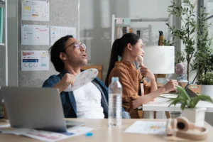 Office workers using handheld fans and looking overheated at their desks in a warm indoor workspace. Best for blogs about offices that are too hot, poor cooling, indoor comfort problems, and commercial AC not keeping up.