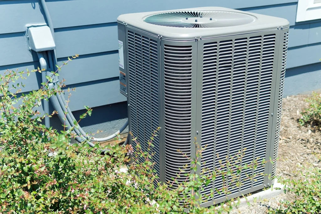 An outdoor air conditioning condenser unit installed against a blue house with connecting refrigerant lines and surrounding vegetation. Best for blogs about AC condenser units, outdoor AC unit maintenance, AC repair, AC replacement, residential cooling systems, signs your outdoor unit has a problem.