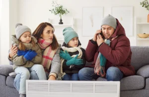 Family dressed in winter clothing and sitting indoors while appearing cold, with one person making a phone call for help. Best for blogs about heating emergencies, furnace failure, no heat, and winter HVAC service.