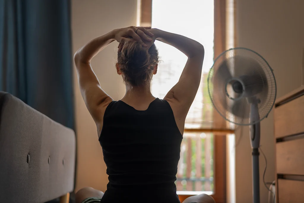 Woman sitting indoors near a portable fan and tying up her hair in a warm room. Best for blogs about summer heat, hot bedrooms, poor cooling, and AC not keeping up.