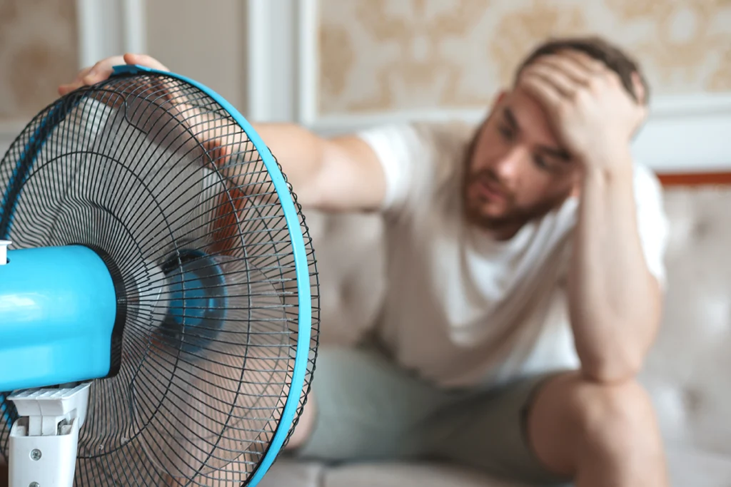 Man sitting indoors beside a portable fan and looking overheated and uncomfortable. Best for blogs about AC not cooling, hot rooms, summer discomfort, and home cooling problems.