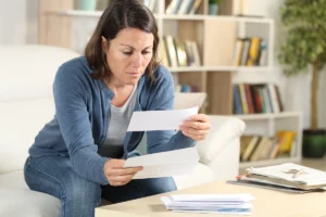 A woman reviewing bills or documents with a worried expression, sitting at a table with stacked papers in a home office setting. Best for blogs about high energy bills, why is my electric bill so high, HVAC energy efficiency, signs your HVAC system needs repair, old HVAC system vs new system savings, and when to replace an inefficient unit.