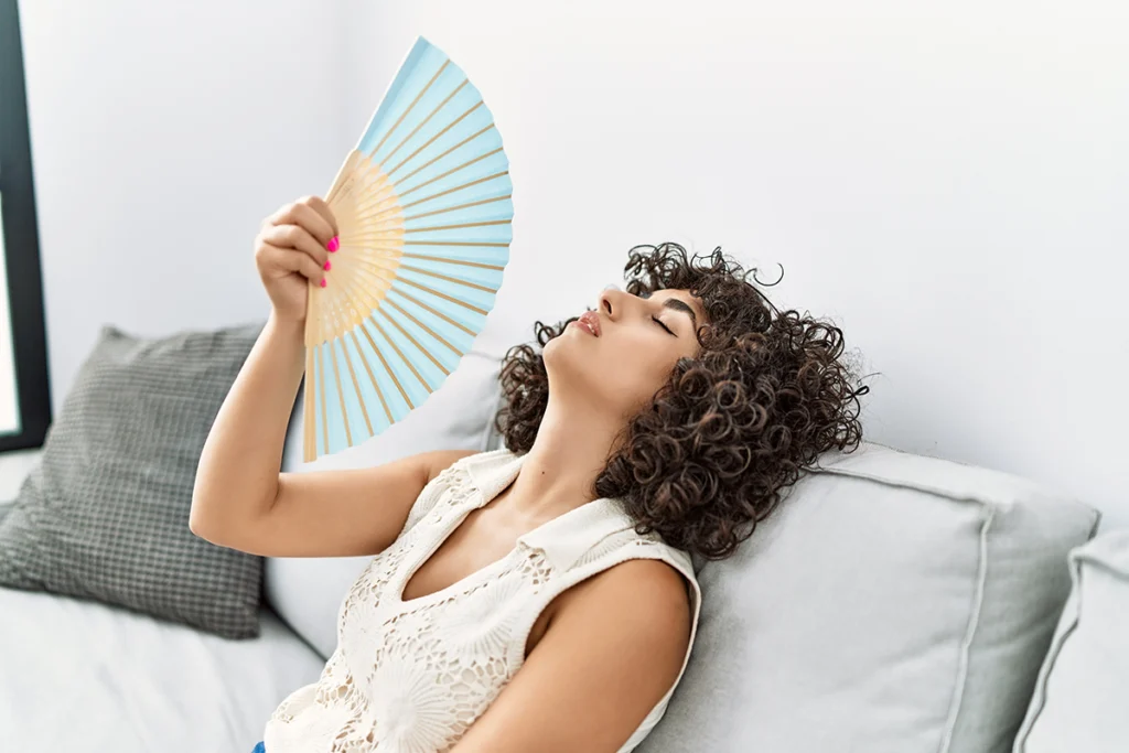 Woman sitting on a couch and fanning herself while appearing overheated indoors. Best for blogs about AC not cooling, summer heat, home discomfort, and indoor cooling problems.