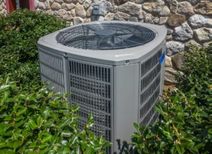 Outdoor residential AC condenser unit surrounded by landscaping near a stone exterior wall.