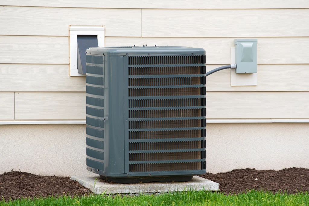 Residential outdoor AC condenser unit installed beside a house with visible electrical disconnect and wall penetration. Best for blogs about air conditioning systems, AC repair, condenser units, and residential cooling equipment.