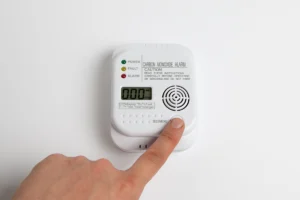 Hand pressing the test button on a wall-mounted carbon monoxide alarm with digital display and indicator lights. Best for blogs about carbon monoxide detector safety, furnace safety tips, gas furnace carbon monoxide risks, signs of carbon monoxide in the home, why every home needs a CO detector, and heating safety during winter.