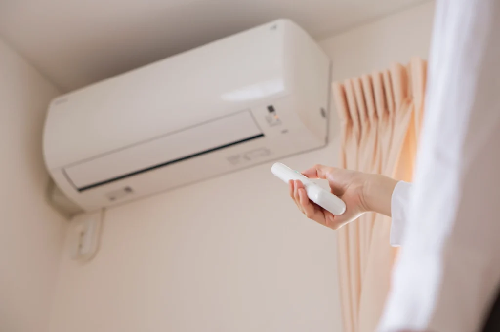 Hand using a remote control to operate a wall-mounted ductless mini split indoor unit. Best for blogs about ductless mini splits, mini split controls, mini split operation, mini split not cooling or heating, zoned comfort, and ductless AC systems.