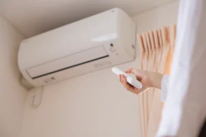 Hand using a remote control to operate a wall-mounted ductless mini split indoor unit. Best for blogs about ductless mini splits, mini split controls, mini split operation, mini split not cooling or heating, zoned comfort, and ductless AC systems.